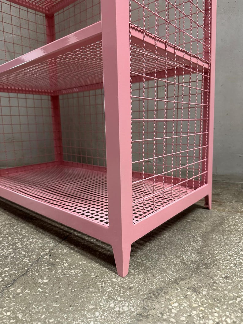 May include: A pink metal storage shelf with three levels. The shelves and sides are made of a wire mesh, and the frame is a solid pink. The legs are angled and the shelf is on a concrete floor.