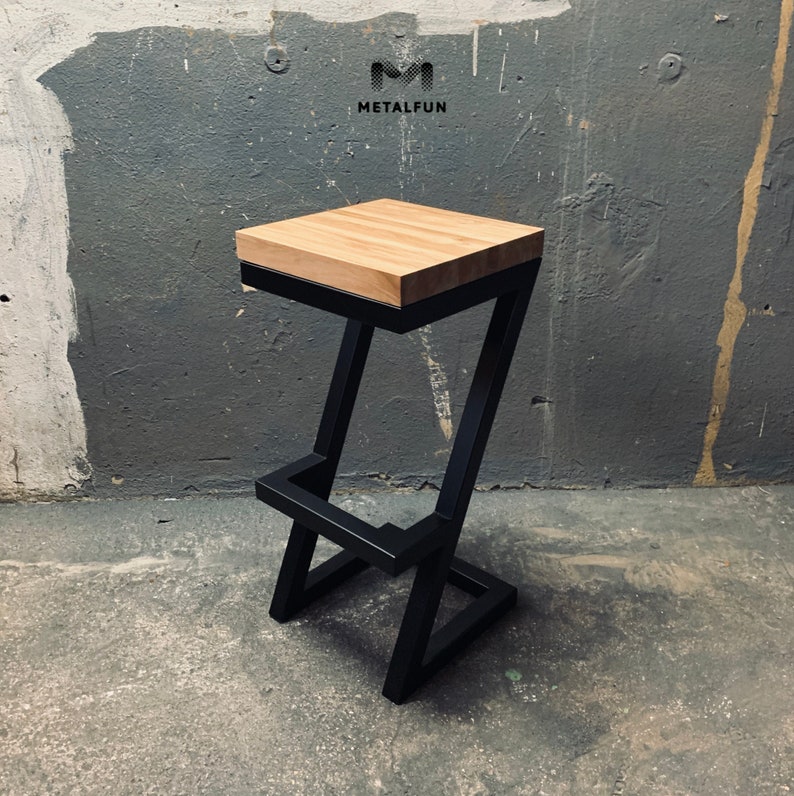 May include: A modern bar stool with a solid wood seat and a black metal frame. The seat is a light brown color, and the frame has a Z-shaped design. The stool is sturdy and stylish, suitable for a kitchen or bar area. The logo "METALFUN" is visible.