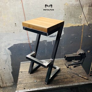 May include: A modern bar stool with a square, light-colored wooden seat. The frame is made of dark metal, with a geometric design. The stool is on a wooden surface. The logo "METALFUN" is visible in the top right corner.