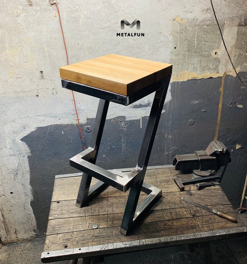 May include: A modern bar stool with a square, light-colored wooden seat. The frame is made of dark metal, with a geometric design. The stool is on a wooden surface. The logo "METALFUN" is visible in the top right corner.