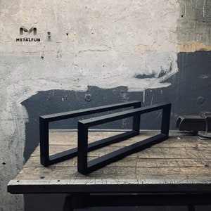 Op de afbeelding: Twee zwarte metalen tafelpoten, elk met een rechthoekig frame, liggen op een houten oppervlak. De poten zijn gemaakt van stevig metaal en hebben een eenvoudig, modern ontwerp.