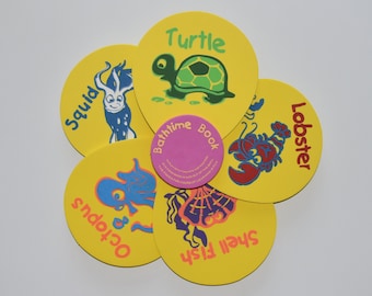 May include: A yellow bath toy set with a flower shape. Each petal features a different sea creature, including a turtle, squid, lobster, octopus, and shell fish. The center of the flower has a pink circle with the text "Bathtime Book".