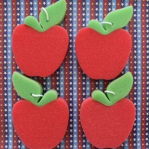 May include: Four red sponge apples with green leaf stems on a blue and red patterned background. The sponges are shaped like apples and have a textured surface.
