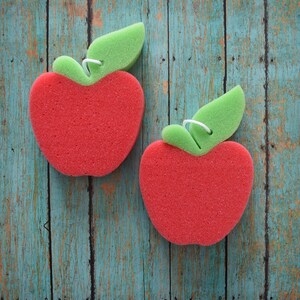 May include: Two red sponge apple-shaped dish scrubbers with green leaf stems on a blue wooden background.