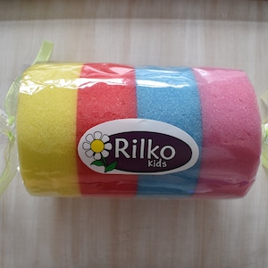 Może przedstawiać: Wielokolorowa gąbka kąpielowa owinięta w przezroczysty plastik z zielono-żółtą wstążką. Gąbka jest żółta, czerwona, niebieska i różowa. Biała etykieta z stokrotką i tekstem "Rilko Kids" znajduje się na gąbce.