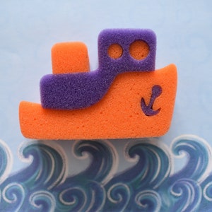 May include: A purple and orange sponge shaped like a boat with an anchor on the side. The boat is sitting on a blue background with a wave pattern.