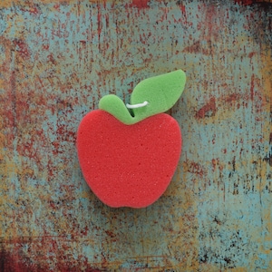 May include: Red sponge in the shape of an apple with a green leaf. The sponge is on a textured blue and brown background.