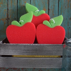 May include: Three red apple-shaped sponges with green leaf accents are displayed in a gray wooden crate. The sponges are arranged in a triangular shape with the largest sponge at the bottom.
