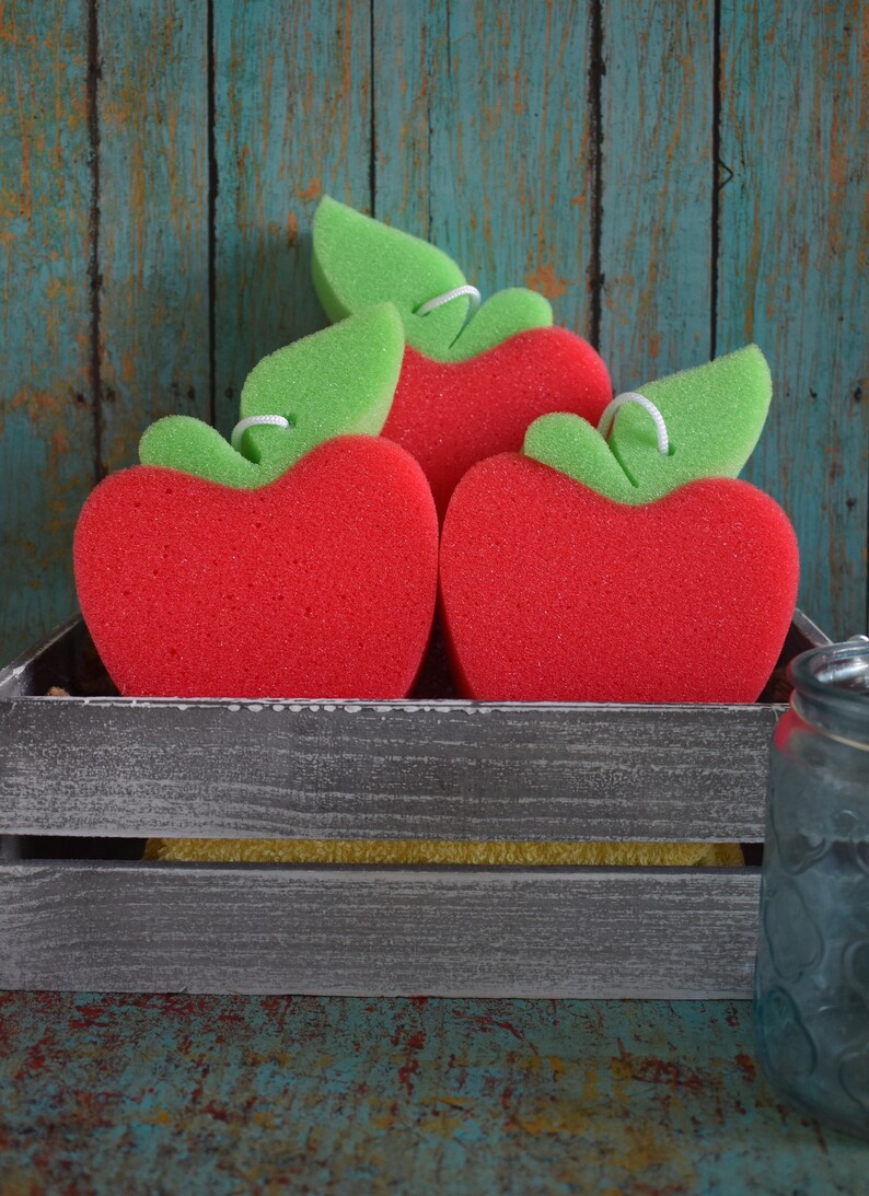 May include: Three red apple-shaped sponges with green leaf accents are displayed in a gray wooden crate. The sponges are arranged in a triangular shape with the largest sponge at the bottom.