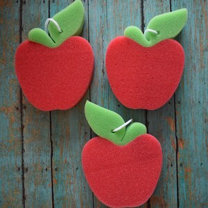 May include: Three red sponge apple-shaped dish scrubbers with green leaf accents. The scrubbers are on a blue and green wooden background.