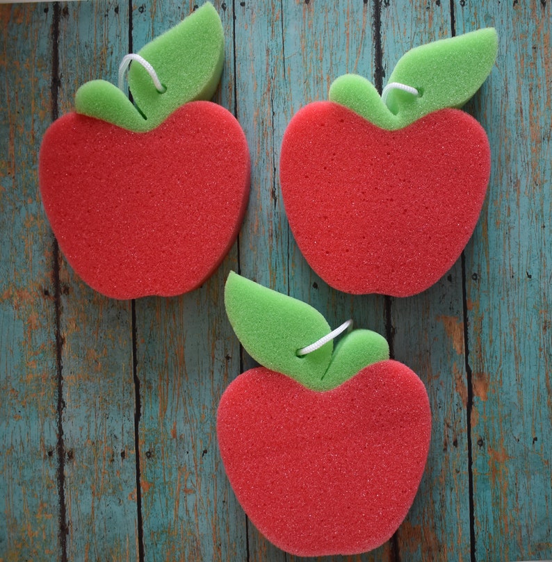 May include: Three red sponge apple-shaped dish scrubbers with green leaf accents. The scrubbers are on a blue and green wooden background.