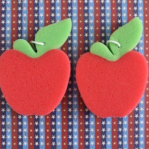 May include: Two red sponge apple-shaped bath scrubbers with green leaf details. The scrubbers are on a blue and red patterned background.
