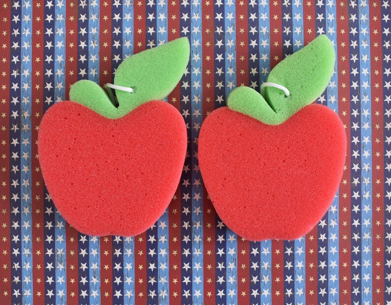 May include: Two red sponge apple-shaped bath scrubbers with green leaf details. The scrubbers are on a blue and red patterned background.