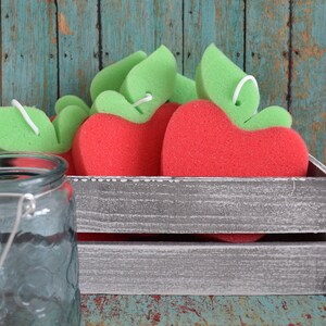 May include: A wooden crate filled with red and green apple-shaped bath sponges. The crate is sitting on a blue and green wooden surface.