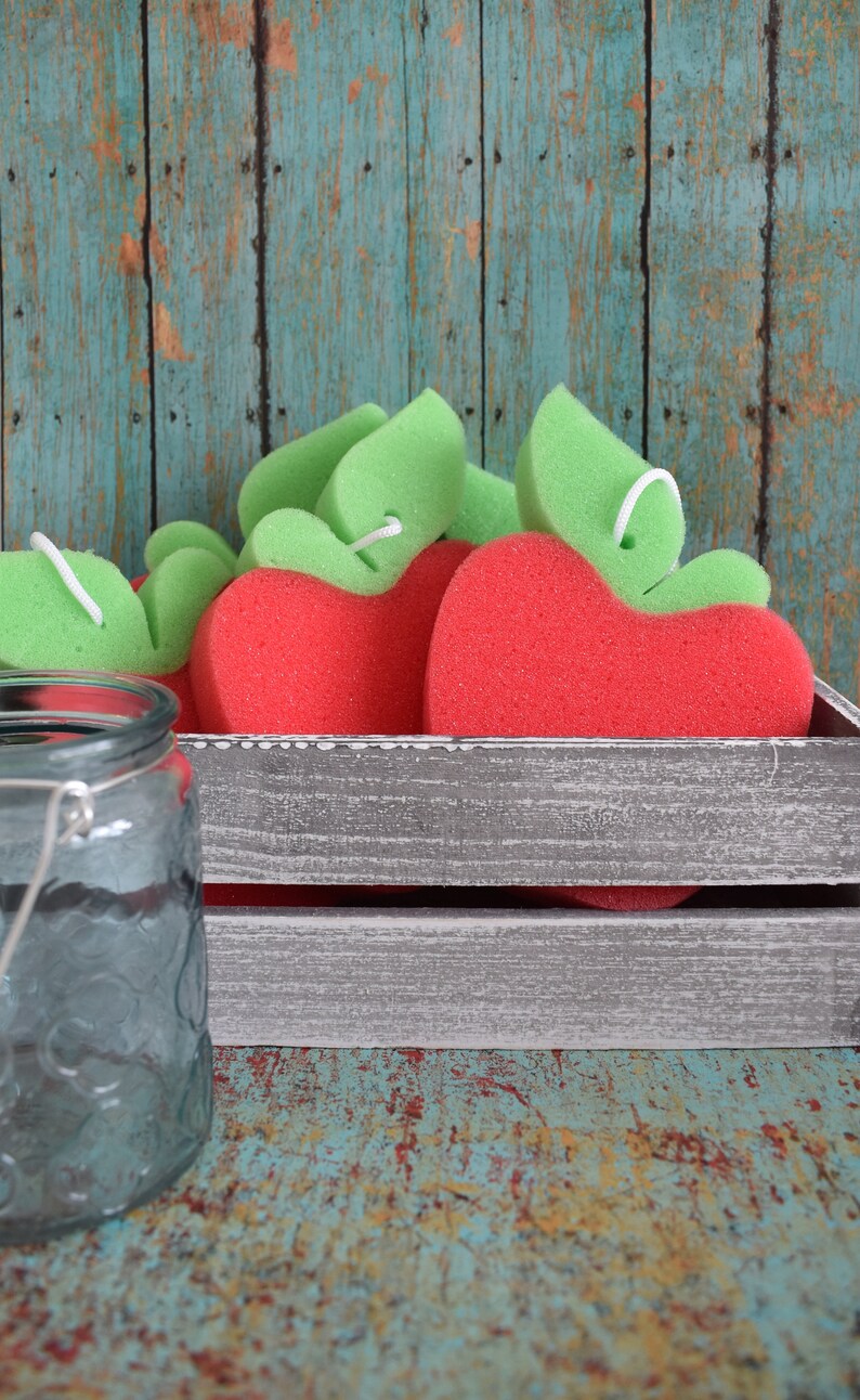 May include: A wooden crate filled with red and green apple-shaped bath sponges. The crate is sitting on a blue and green wooden surface.