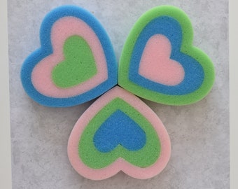 May include: Three heart-shaped sponges in shades of pink, green, and blue. Each sponge has a concentric circle design with a smaller heart inside.