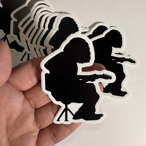 May include: A collection of black and white stickers featuring a silhouette of a person playing a guitar while seated on a stool. The stickers have a white border and are stacked on top of each other.