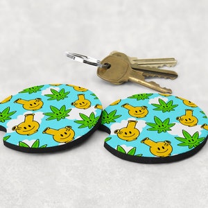 May include: Two round car coasters with a blue background, featuring a pattern of yellow smiling bongs, green cannabis leaves, and white clouds. A set of keys rests above the coasters.