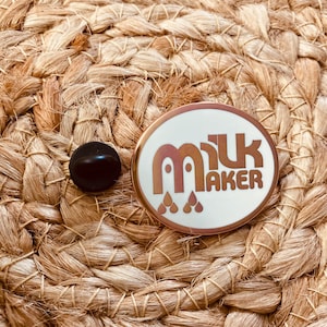 May include: A white enamel pin with a rose gold border and a brown graphic that says "milk maker" with three stylized drops of milk underneath the text.