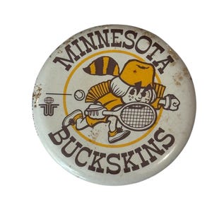 May include: A round white pinback button featuring the brown text MINNESOTA BUCKSKINS. The centre graphic depicts a cartoon character wearing a yellow hat and shirt, holding a tennis racket, with a small raccoon tail and a logo to the left.