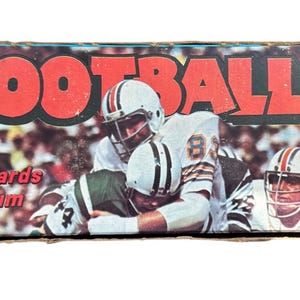 Könnte beinhalten: Eine rechteckige Pro Football Sammelkarten- und Kaugummischachtel mit einer Grafik von Footballspielern in weißen Helmen und Trikots. Der Text lautet Pro Football und 15 cents, vor einem unscharfen Stadionhintergrund.