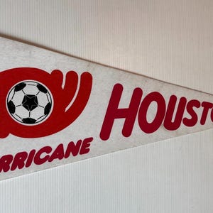 Puede incluir: Un banderín triangular con las palabras "HOUSTON HURRICANE" en rojo, y un gráfico de balón de fútbol. El banderín tiene un borde naranja y el logotipo de la NASL.