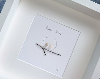 May include: A white framed picture with a white background and the text "Love birds..." written in black ink. Two small white birds are perched on a twig, facing each other.