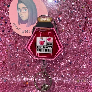 May include: Pink love potion bottle shaped retractable badge holder with a clear plastic cover and a silver spring clip. The bottle has a black lid and a label that says "Love Potion" and "XOXO".