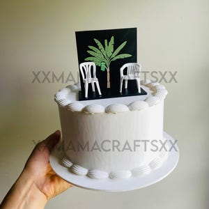 May include: A white frosted cake topped with a black rectangular cake topper featuring a green banana tree illustration and two miniature white plastic chairs. The cake has a smooth white finish with piped frosting borders along the top and base.
