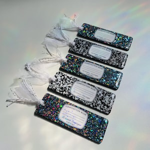 May include: A collection of composition book-themed bookmarks. The bookmarks feature a glittery, speckled design in various colors, with a white label that reads "COMPOSITION BOOK". Each bookmark has a white ribbon attached for easy use.