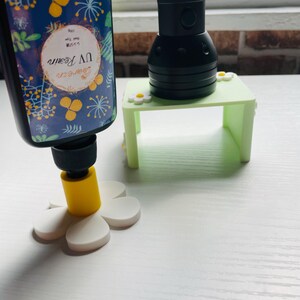 May include: A black bottle of UV resin with a blue label featuring yellow floral patterns, standing on a white flower-shaped base with a yellow center. Next to it is a black UV flashlight resting on a small, light green rectangular stand.