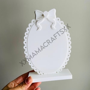 May include: A white, oval-shaped decorative sign with a scalloped edge and a bow at the top. The sign is mounted on a rectangular base. The text "XXMAMACRAFTSXX" is printed on the sign.
