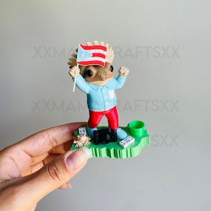 May include: A figurine of a coqui frog wearing a light blue shirt and red pants, holding a Puerto Rican flag. The figure stands on a green base with a smaller frog, a black camera, two white dominoes, and a small green cup accessory included.