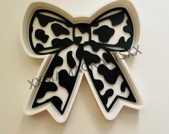 May include: A white bow-shaped cookie cutter with a black cow print design. The bow has a detailed outline and is set against a white background. The design is suitable for baking or crafting.