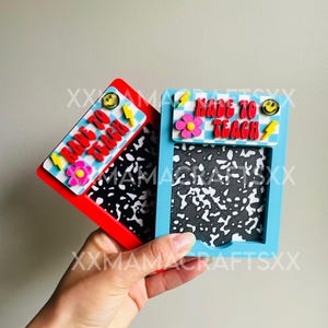 May include: Two decorative notebook-style frames, one red and one blue, held in a hand. Each frame has the text "MADE TO TEACH" with a flower, lightning bolt, and smiley face. The frames have a black and white speckled pattern.