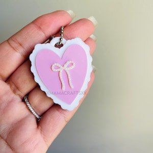 May include: A pink heart-shaped keychain with a white scalloped border and a small bow detail. The keychain has a silver chain and the text "XMAMACRAFTSXX" is visible on the heart.