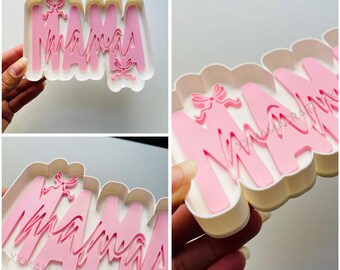 May include: A pink "Mama" cookie cutter with a bow design. The letters are in a cursive font and set in a white scalloped frame. The image is a close-up shot of the cookie cutter.