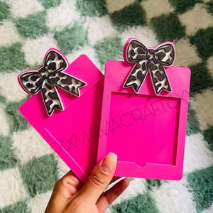 May include: Two bright pink photo holders with a bow-shaped embellishment. The bows have a black and white spotted pattern. The text "XXMAMACRAFT" is visible on the photo holder. The photo holders are held in a hand.