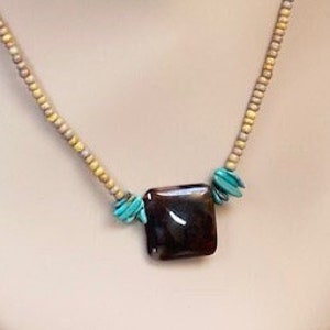 May include: A brown and black square pendant hangs from a beige and yellow beaded necklace. The pendant is accented with turquoise beads.