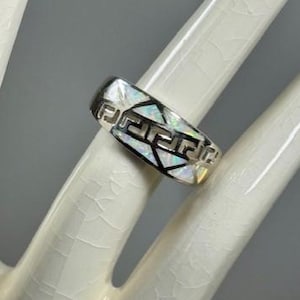 May include: A silver ring with a geometric design and iridescent inlay. The ring features a Greek key pattern and is displayed on a white hand.