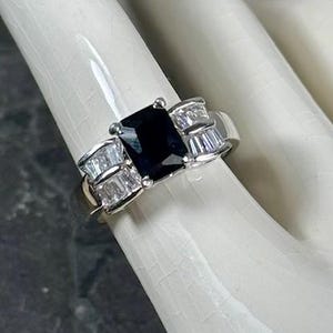 May include: A silver ring with a black square-shaped gemstone surrounded by smaller clear gemstones.