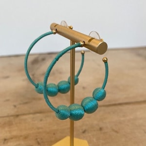 May include: A pair of turquoise hoop earrings with three turquoise beads. The earrings are displayed on a gold-colored earring stand with a hexagonal base. The earrings are made of a woven material.