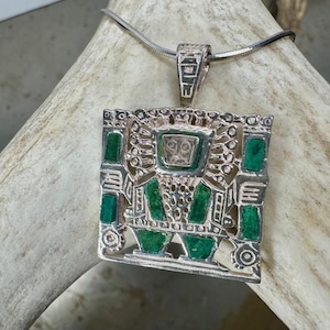 May include: A silver-toned pendant with a square, openwork design. The pendant features rectangular green insets and a central square element. It hangs from a thin, silver-colored chain. The pendant has an intricate, geometric pattern.