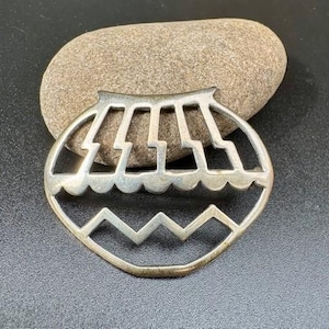 May include: A silver-colored, openwork metal pendant with a Southwestern design. The pendant features a stylized mountain range at the bottom and a series of rectangular shapes at the top. The pendant is resting on a smooth, oval-shaped stone.