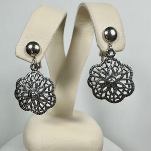 May include: Silver-toned drop earrings with a floral filigree design. Each earring features a small, round stud and a dangling, openwork flower shape. The earrings are displayed on a cream-colored jewelry stand.