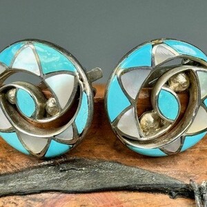 May include: A pair of silver earrings with turquoise and mother-of-pearl inlay. The earrings are round and have a geometric design.
