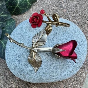 May include: A decorative metal rose brooch with a red flower and gold-colored leaves and stem. The brooch is designed to resemble a realistic rose and is set against a gray stone background.