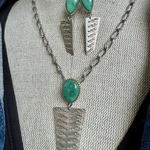 May include: A silver-toned jewelry set consisting of a necklace and matching earrings. Each piece features a teardrop-shaped green stone set above a textured, trapezoidal silver-toned metal pendant with a repeating geometric pattern.
