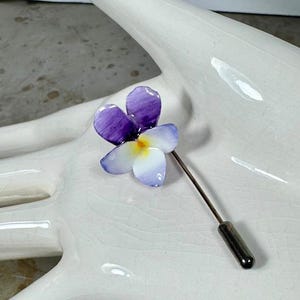May include: A floral lapel pin featuring a flower with purple and white petals and a yellow center. The pin has a long, slender, silver-toned stem and a dark gray cap. The flower is set against a white background.