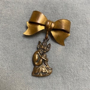 May include: A gold-toned metal brooch featuring a small, detailed sculpture of a fairy sitting on a mushroom, with a large bow attached above.